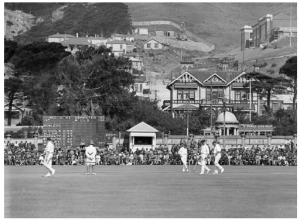 The photograph was taken at the Basin Reserve in January 1930. It was a great day for New Zealand cricket. Batting first the New Zealand openers put on 276 for the first wicket, for many years a New Zealand record, and the scoreboard shows the moment when both openers reached their centuries. In the background above the Basin work is just beginning on the car tunnel, while to the right the north wing of the Wellington Girls’ College building hall is nearly complete. Outside the ground a tram advertising soap rumbles along Dufferin Street, between the Wakefield Memorial and the long-demolished houses on the other side. The scoreboard and the little scorers’ shed in the northeast corner had been erected a few years before. Nowadays the scoreboard is on the southern side. (Photo: G-2-¼-EP, Alexander Turnbull Library). 